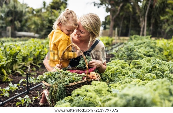 Bonding in the garden. Happy young mother smiling\
cheerfully while carrying her daughter and picking fresh kale in a\
vegetable garden. Self-sufficient family gathering fresh produce on\
their farm.