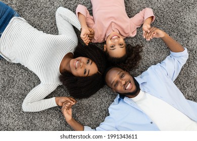 Bonding And Lifestyle Concept. Above Top High Angle View Portrait Of Black Family Of Three People Lying On Floor Rug Carpet Together At Home Indoors. Happy Parents And Child Smiling, Holding Hands