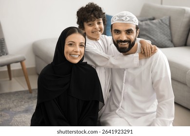 Bonding Muslim family portrait. Happy friendly Middle Eastern couple in traditional attire with schoolboy child son cuddle at living room united with love care Islamic values look at camera with smile - Powered by Shutterstock - Get 15% off with code: PIKWIZARD15