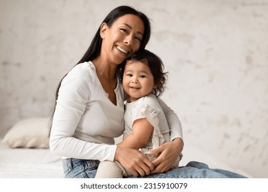 Bonds Of Love. Happy Asian Mom And Baby Daughter Sharing Warm Embrace, Posing Smiling To Camera Sitting On Bed Together At Home. Joyful Moments Of Parenthood And Babyhood Concept - Powered by Shutterstock