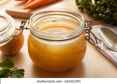 Bone broth made from chicken in a glass jar, with carrots, onions, and parsley in the background