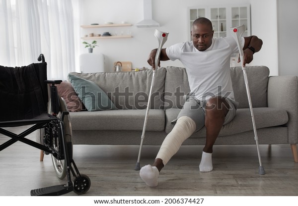 Bone fracture after incident, treatment at home,\
recovery. Black middle aged male with broken leg with plaster cast\
gets up from sofa with crutches in living room interior with\
wheelchair, empty space
