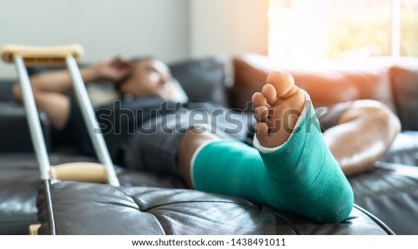 Bone fracture foot and leg on male\
patient with splint cast and crutches during surgery rehabilitation\
and orthopaedic recovery lying on couch staying at\
home