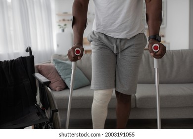 Bone Fracture, Injury, Recovery And Treatment At Home After Incident. Mature Black Man With Broken Leg With Plaster Of Cast Walk With Crutches In Living Room Interior With Wheelchair, Cropped