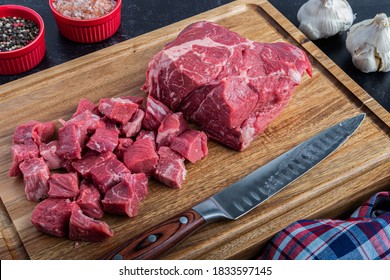 A boneless beef chuck roast being cubed on a cutting board with sea salt and peppercorns next to a carving knife