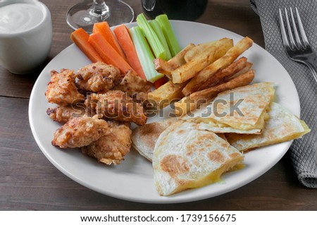Boneless chicken sampler with flour tortillas quesadilla fries, celery and carrot sticks on a white plate and ranch dip on the side
