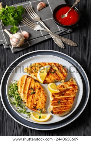 boneless skinless juicy grilled chicken cutlets with tomato sauce and lemon slices on plate on black wooden table with forks, vertical view from above