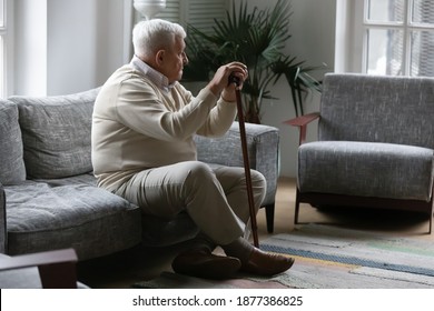 Bones ache on weather. Thoughtful mature male with grey hair resting on sofa leaning on stick feeling hard to move. Sad tired old man suffer of walking with limp need rehabilitation after leg trauma