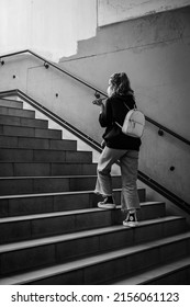 BONN, GERMANY - Apr 26, 2022: A Girl In A Medical Mask With A Backpack , Talking On The Phone , Walking Up Subway Stairs Grayscale Shot