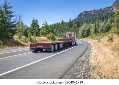 Bonnet Powerful Yellow Big Rig Semi Truck With Empty Flat Bed Semi Trailer Running On The Winding Highway Road With Trees And Mountain In Columbia Gorge Area To Warehouse For Pick Up Next Load