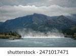 The Bonneville Dam is releasing water into the Columbia River in the midst of a mountainous landscape. Low clouds hang close to the forested peaks, adding a mystic atmosphere to the scene.