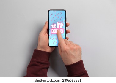 Bonus gaining. Man using smartphone on light grey background, top view. Illustration of open gift boxes, word and falling confetti on device screen