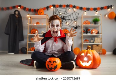 Boo, scare on Halloween. Portrait of terrifying little boy in traditional Halloween vampire costume who screams and scares looking at camera. Child sits on floor near pumpkin in room with festive deor