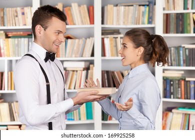 Book Is The Best Gift. Cheerful Young Nerd Man Giving A Book To A Beautiful Young Woman While Standing At The Library