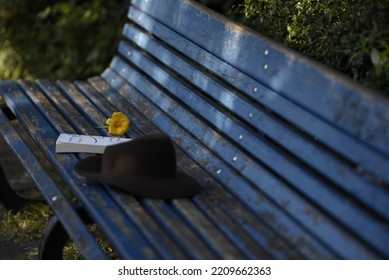 A Book Hat And A Yellow Flower On A Blue Bench. A Reader's Props Left On A Bench In The Park. Romantic Photo Of A Blue Wooden Bench And Objects On It.