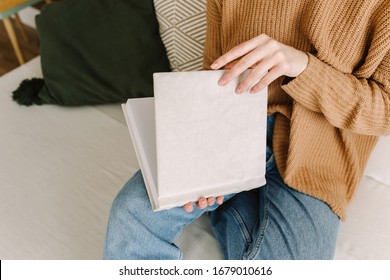 Book mock-up in hands of a woman. Album with clean cover. Book with a cover of cream textile. Empty book template. Young woman holds the empty book in her hands close-up. Girl is sitting on the couch
