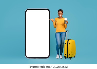Booking App. Happy Black Tourist Lady Withn Suitcase Pointing At Big Blank Smartphone, Excited African American Female Traveller Advertising Mobile Application For Buying Tickets Online, Mockup