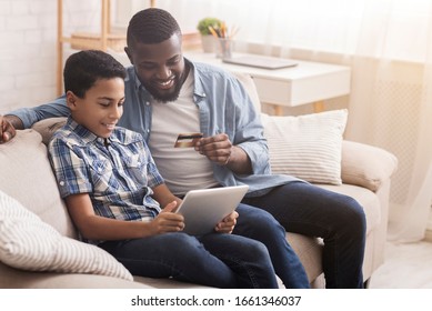 Booking Sport Tickets Online. Portrait Of Happy Afro Father And His Teen Son With Credit Card And Digital Tablet At Home