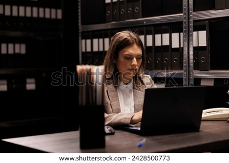 Bookkeeper analyzing administrative files on laptop computer, searching for bureaucracy record in corporate depository. Manager in formal suit working at accountancy report in storage room