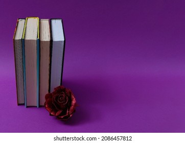 Books set upright, in front of them a red glittering rose on a violet background. Text space. Book lovers, mother's day, holiday season, gift concept. Minimal style.