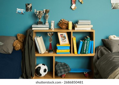 Bookshelf standing between two beds holding trophies, medals, books, framed photo, baseball glove, and toy van with soccer ball, skateboard, and teddy bear placed nearby on floor