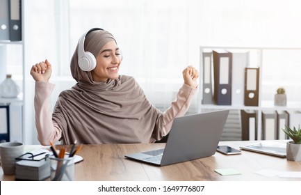 Boost Mood At Work. Joyful Arabic Woman In Hijab Listening Music In Wireless Headphones In Office And Dancing While Sitting At Desk, Panorama