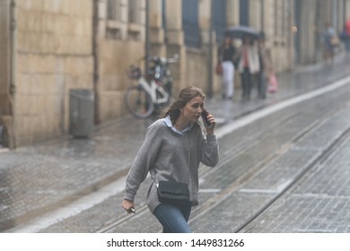 Bordeaux / France - June 21, 2019: Lifestyle Of Big City In Rainy Summer Day. Young Girl Without Umbrella Talking On A Smartphone And Running Through The Tram Tracks In The Rain. High Resolution Image