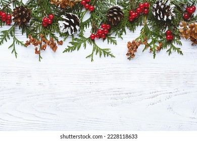 Border of Christmas tree branches pine cones flowers and red berries on white wooden background, copy space