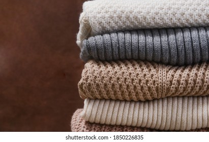 Border Of Pile Of Knitted Woolen Sweaters Autumn Colors. Clothes With Different Knitting Patterns Folded In Stack. On Brown Linen Background, Warm Cozy Winter Fall Knitwear Concept 

