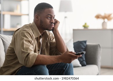 Bored African American Man Watching Boring TV Switching Channels Holding Television Remote Controller Sitting On Sofa At Home. Side View Shot Of Discontented Televiewer