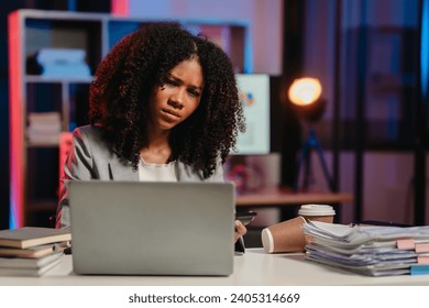 bored African American office worker with an afro, resting her cheek on her hand while looking at a laptop screen.