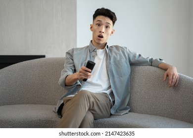 Bored Asian Man Turns On TV With Remote Control To Watch Interesting Program. Guy Sits On Soft Couch Getting Surprised After Changing TV Channel