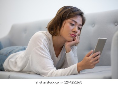 Bored Asian Phone User Browsing Internet. Young Woman In Casual Lying On Couch, Leaning Cheek On Hand And Using Smartphone. Boredom Concept