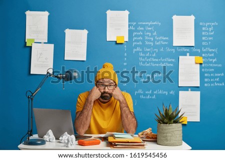 Bored bearded man works with laptop device and papers, holds cheeks, looks at camera, prepares for studying seminar, has deep knowledge of programming, isolated over blue wall with sticky notes