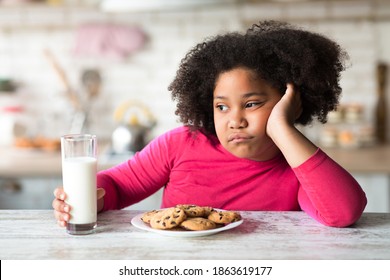 Bored Black Little Girl Sitting At Table With Milk And Cookies, Refuse To Eat Breakfast, Unhappy Kid Resting Head On Hand And Looking Away, Naughty Child Without Appetite Sitting In Kitchen, Closeup