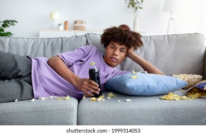 Bored Black Teenager Lying On Couch With Scattered Food, Watching Dull Show Or Movie On TV, Killing Time At Home. African American Adolescent Being Lazy, Eating Snacks, Staring Into Television