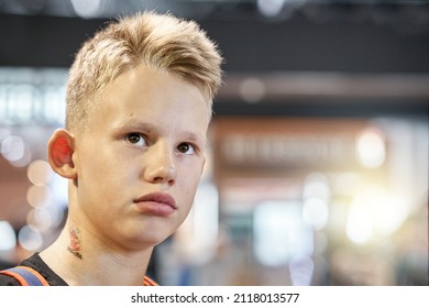 Bored Blond-haired Teenage Boy With Serious Face Expression Sits In Airport Waiting Hall Against Blurred Crowd Extreme Closeup