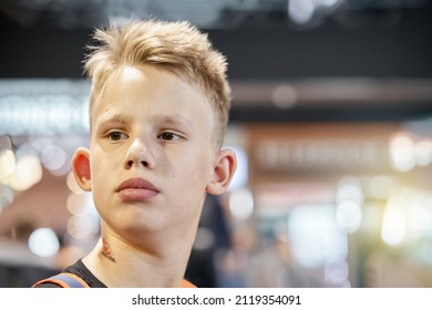 Bored Blond-haired Teenage Boy With Serious Face Expression Sits In Airport Waiting Hall Against Blurred Crowd Extreme Closeup