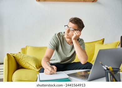 bored charming man with glasses in yellow couch at home writing in notes and looking to window