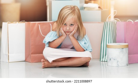 Bored Child, Fashion And Shopping Bags Waiting On Floor In Kids Clothing Shop, Store Or Boutique With Cute Girl Sitting Looking Upset. Portrait Of Kid Shopper Or Customer With Sale Purchase Packages