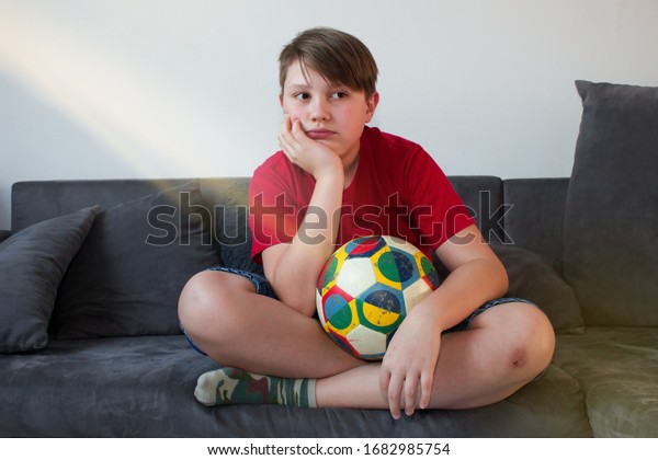 Bored child\
sitting on the couch and holding a football ball.Stay at home.\
Teenage boy bored sitting with kicking ball on couch. Quarantine\
due to coronavirus\
pandemic.