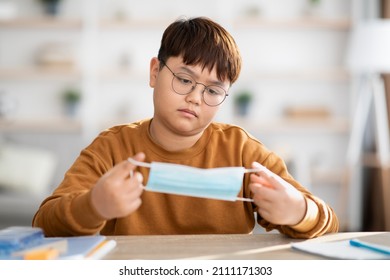 Bored Chubby Asian Teen Boy With Glasses Sitting At Table With Books And Notepads, Holding Protective Face Mask, Tired Of Home Schooling While COVID-19 Pandemic, Copy Space, Home Interior