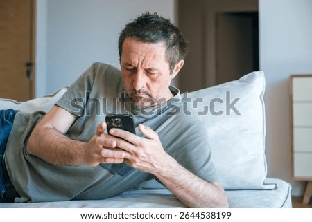 Bored couch potato guy using mobile phone while laying on living room sofa, selective focus