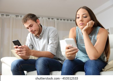 Bored Couple Online With Their Smart Phones Sitting On A Couch In The Living Room At Home