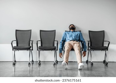 Bored and disappointed male indian employee sitting in waiting room on row of chairs, feeling tired and upset, full length shot, free space