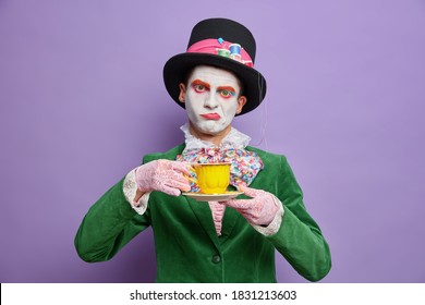 Bored displeased man hatter obsessed with drinking tea wears big hat green costume bowtie poses against purple background comes on spooky party celebrates halloween stands indoor. Fictional character