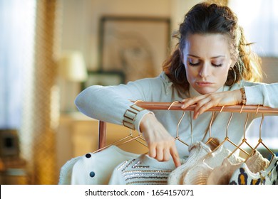 Bored Elegant Middle Age Woman In White Sweater And Skirt In The Modern House In Sunny Winter Day Choosing Sweaters Hanging On Copper Clothes Rack And Doesn't Find Something Suitable.