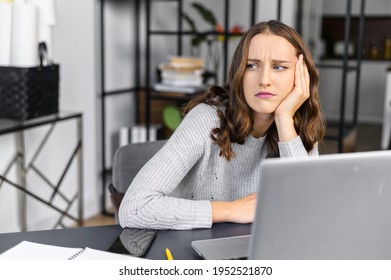 Bored Female Office Employee Sits At The Desk In Front Of A Laptop In The Office, Looks Away And Feels Sad, A Pensive Young Woman Does Not Have Inspiration For Work, Feels Lack Of Sleep And Fatigue