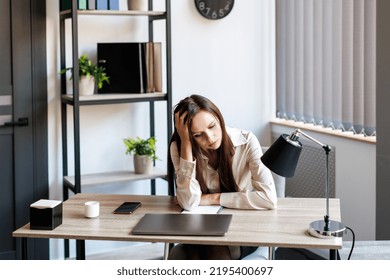 Bored Female Office Employee Sits At The Desk In Front Of A Laptop In The Office, Feels Sad, A Pensive Young Woman Does Not Have Inspiration For Work. Overwork, Working Overtime And Stress At Work