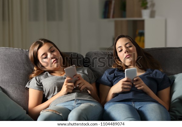 Bored friends using their smart phones\
sitting on a couch in the living room at\
home